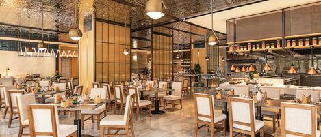 A spacious restaurant with wooden chairs and tables, a high ceiling with hanging lights, and a kitchen area visible in the background.