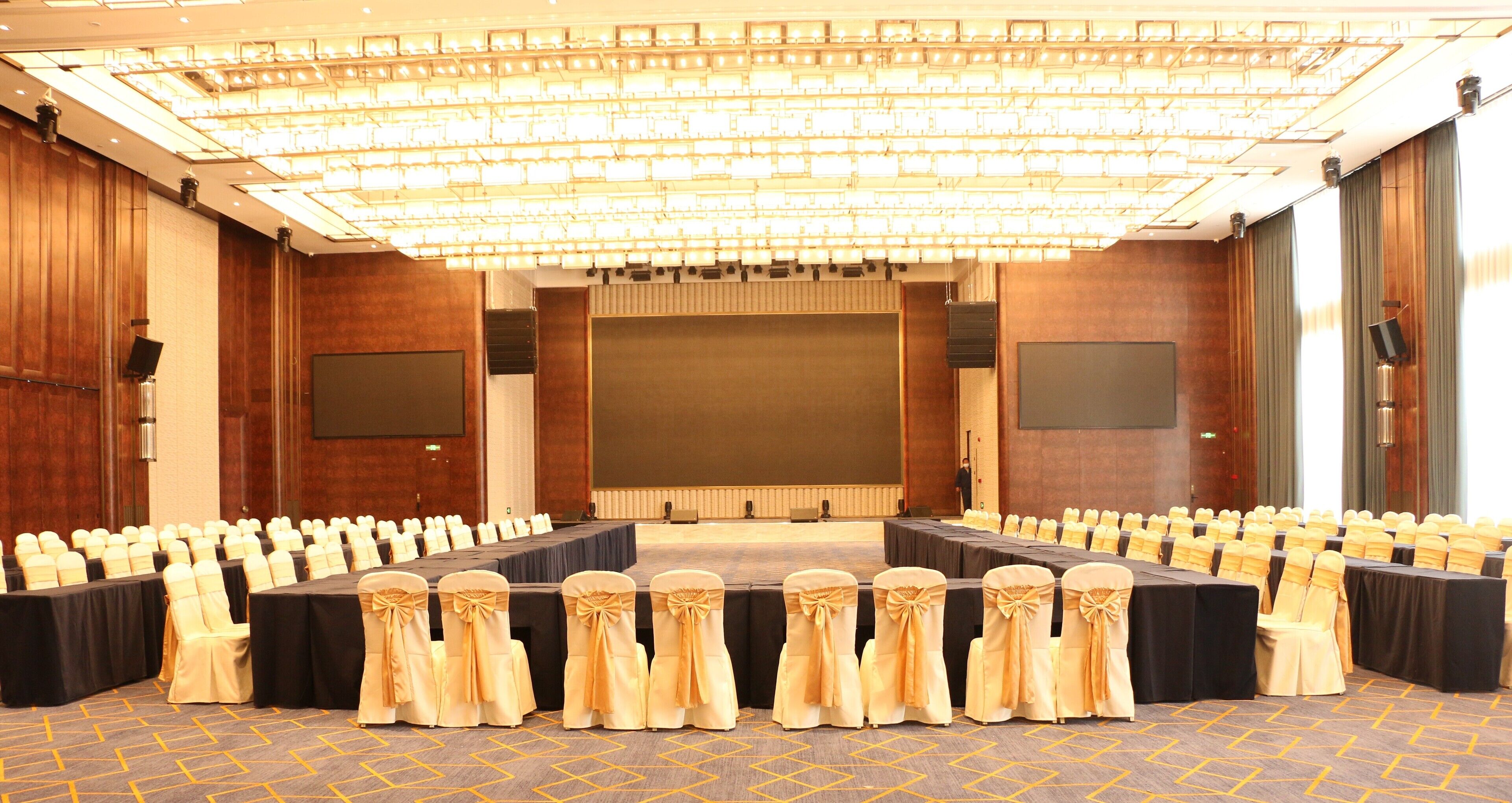 Une salle de conférence avec des rangées de tables et de chaises, un grand écran et une scène.
