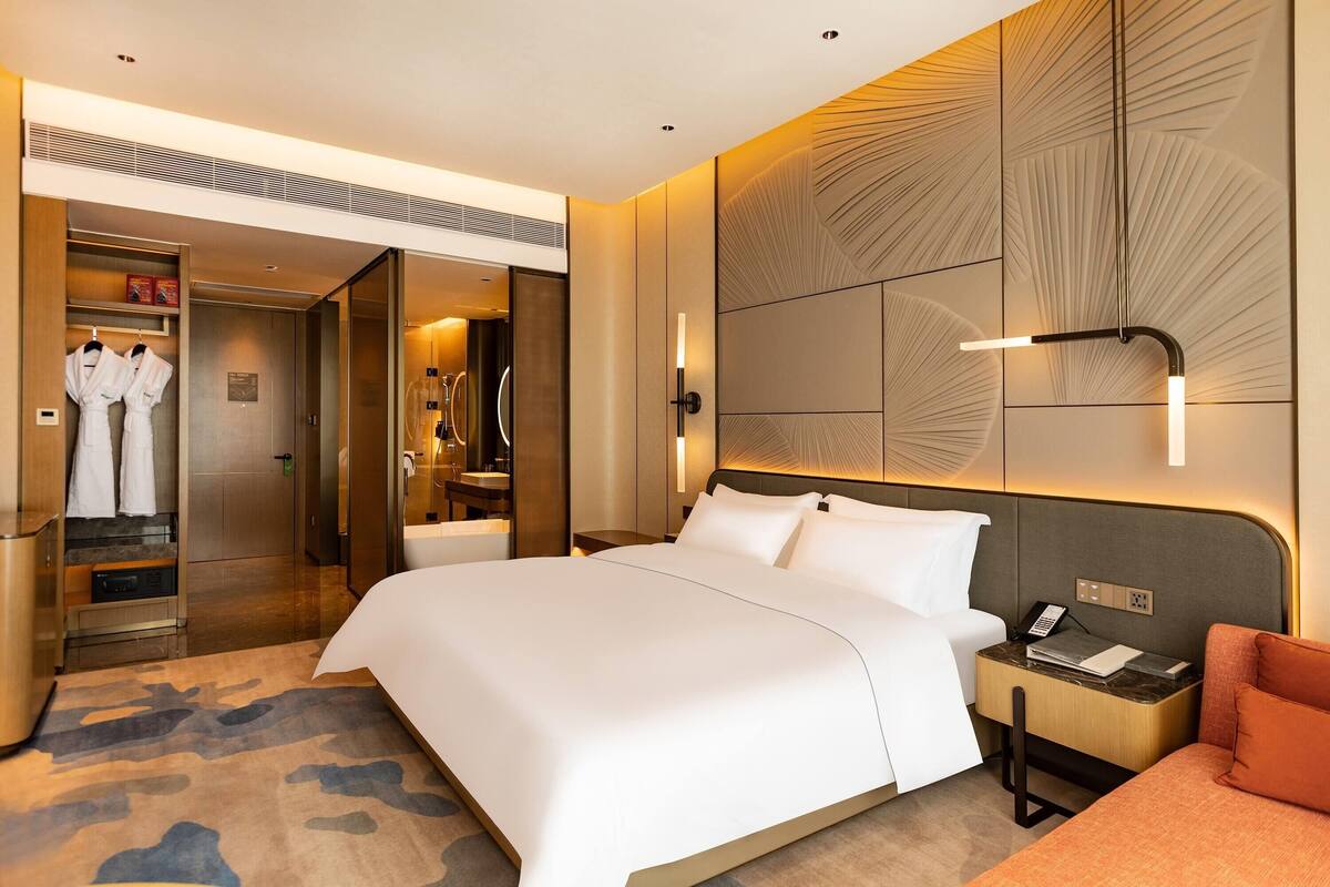 Une chambre d’hôtel moderne avec un grand lit, une table de chevet avec une lampe, une armoire et une salle de bain visible à travers une porte ouverte.