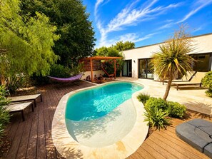 Piscina com deck de madeira, rodeada por vegetação e uma pérgula.