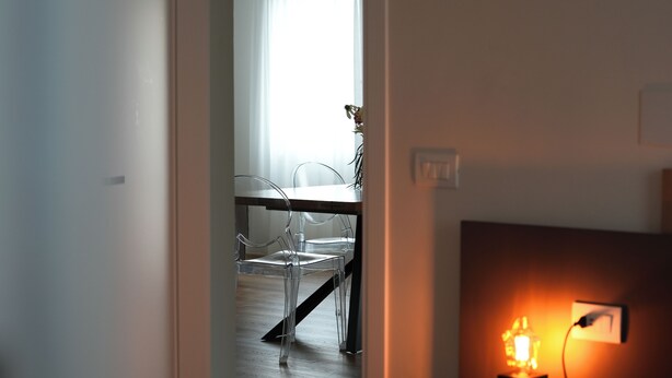 Een hotelkamer met een bed, nachtkastje met lamp, een stoel, en een tafel met stoelen op de achtergrond.