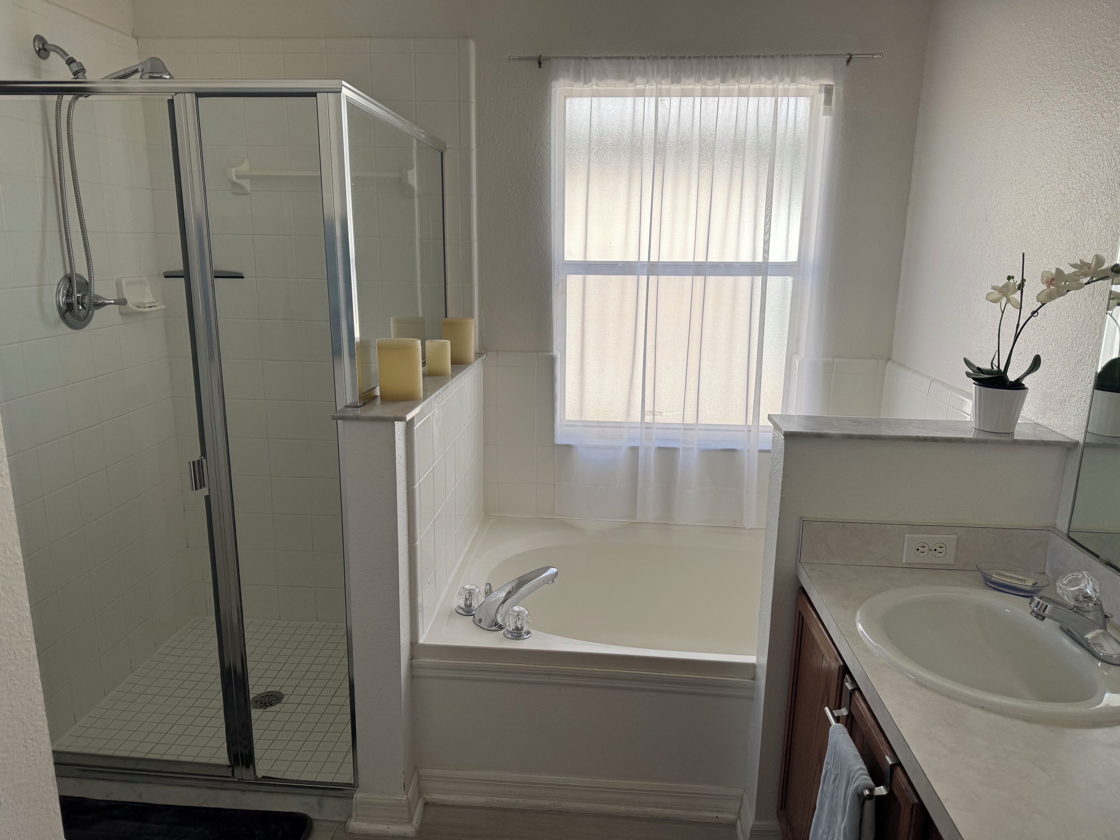 A bathroom with a glass-enclosed shower, a bathtub, a sink, and a window with sheer curtains.