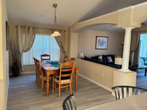 Un coin repas avec une table et des chaises en bois, un luminaire suspendu et une vue sur un salon comprenant un canapé et une lampe.