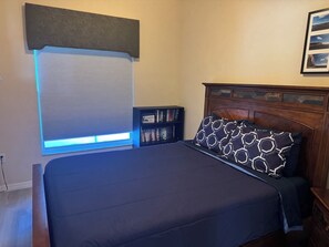 Une chambre avec un grand lit, une bibliothèque, une fenêtre avec des stores et un tableau encadré au mur.