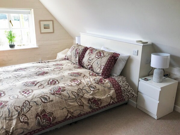 A bedroom with a bed, a nightstand, a lamp, a window with a plant, and a framed picture on the wall. - 1 bedroom accommodation in Wellow, near Yarmouth (Wellow, near Yarmouth)