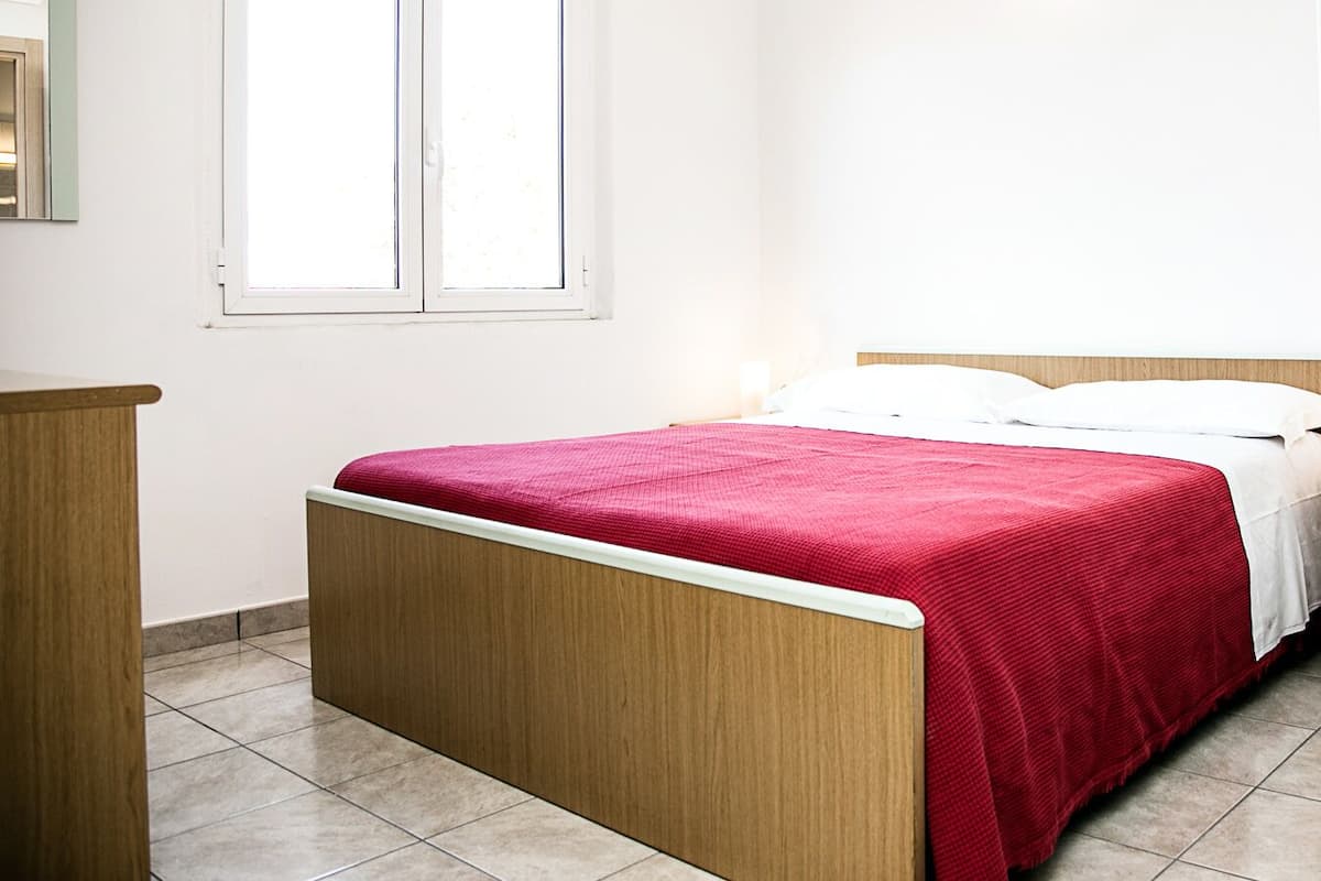 Un dormitorio con estructura de cama de madera, colcha roja, almohadas blancas y una ventana con persianas.