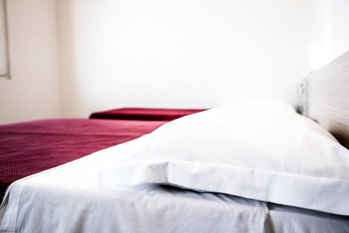 Una cama con ropa de cama blanca y roja, una pared lisa y una ventana al fondo.