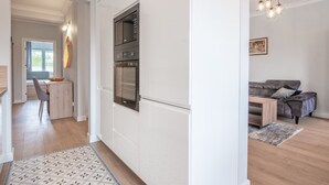A modern kitchen with white cabinetry, a built-in oven, and a microwave. There is a dining area with a table and chairs, and a gray sofa in the living room.