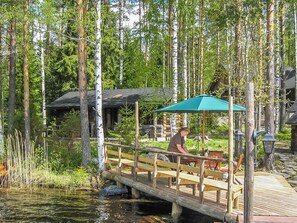 A wooden deck over water with a person seated under a green umbrella, surrounded by trees. - Kaidan kunkku by Interhome (Ristiina)