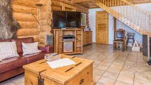 Interior de una cabaña de troncos con escalera de madera, televisión, sofá y una mesa de centro.