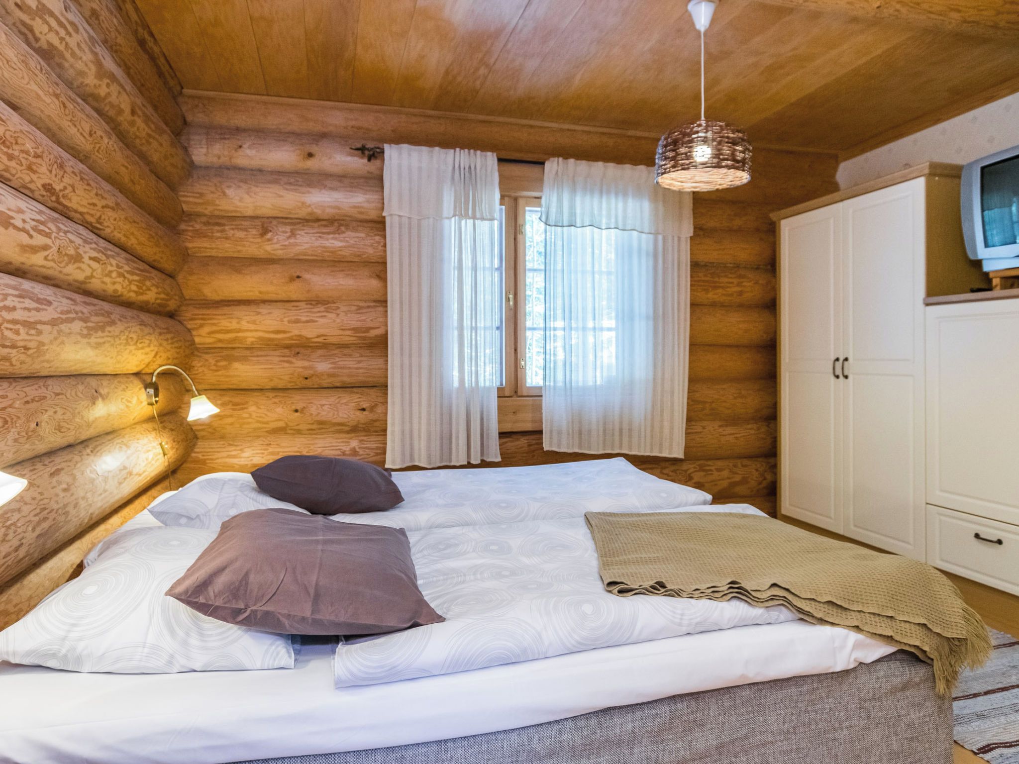 A log cabin bedroom with a bed, pillows, a hanging light fixture, a wardrobe, and a window with curtains.