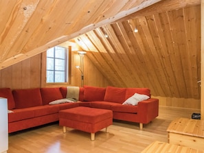 A cozy attic room with a wooden sloped ceiling, a sectional red sofa, a small wooden table, and a floor lamp.