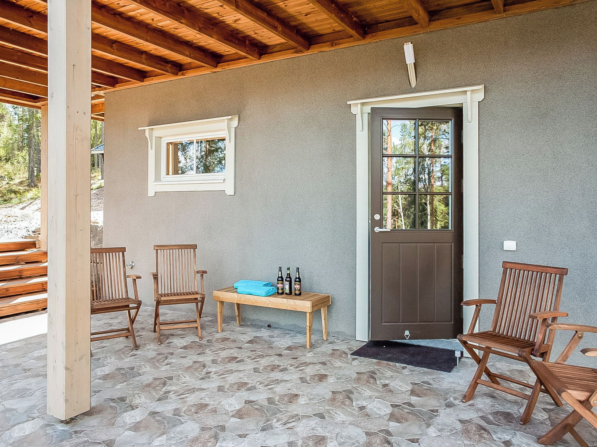 A covered outdoor seating area with stone flooring, wooden chairs, a bench, and a door with a window.