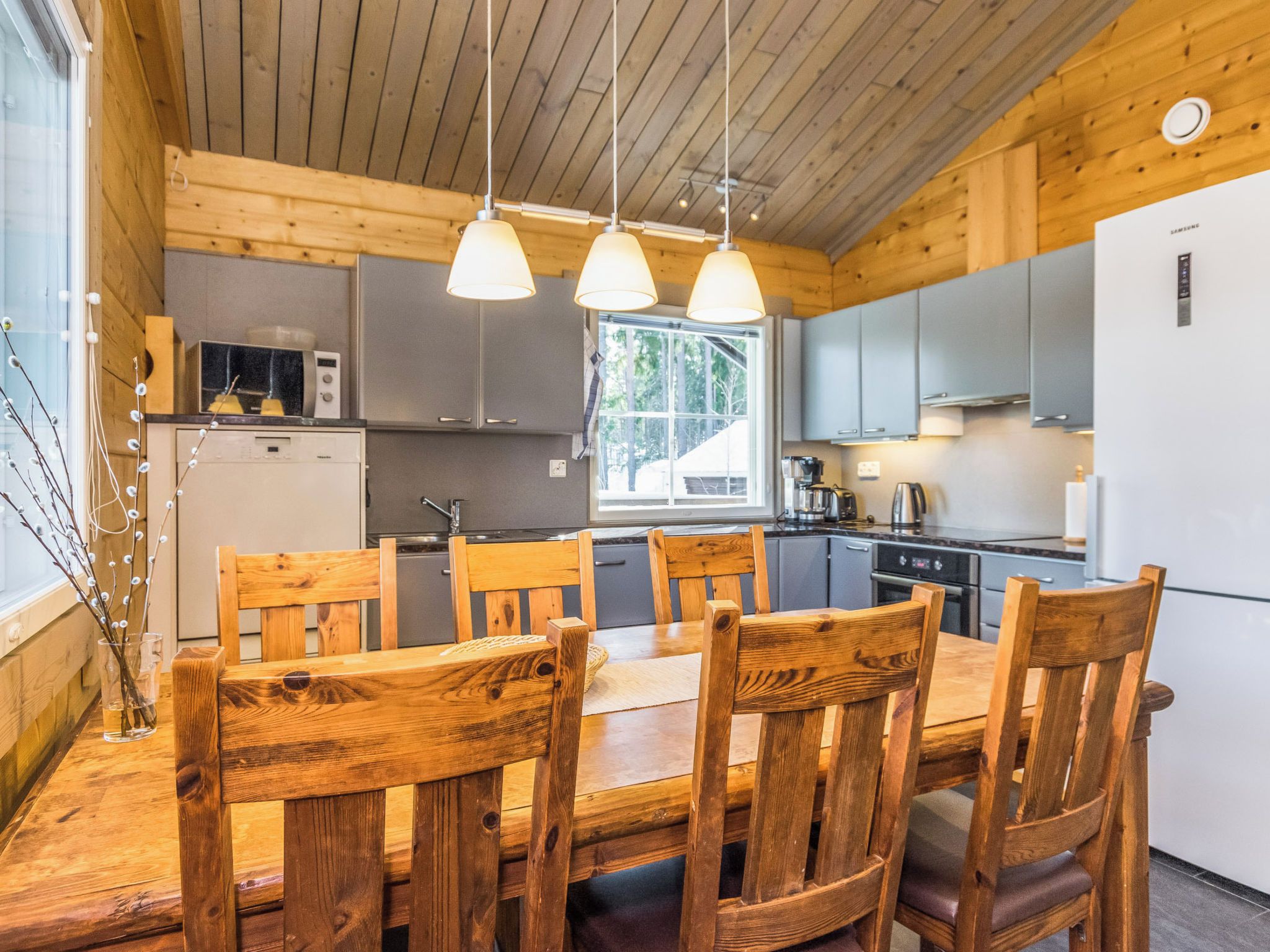 Interior de madera con mesa y sillas de comedor, electrodomésticos de cocina e iluminación colgante.