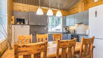 Interior de madera con mesa y sillas de comedor, electrodomésticos de cocina e iluminación colgante.