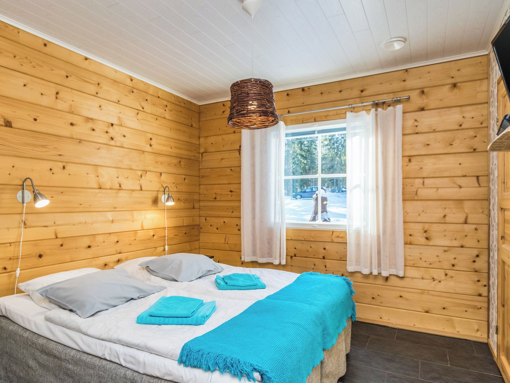 Une chambre dans un chalet en bois, avec un lit, deux lampes de chevet, une fenêtre avec des rideaux et un luminaire suspendu.
