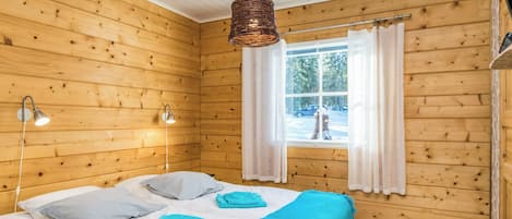 A wooden cabin room with a bed, two bedside lamps, a window with curtains, and a hanging light fixture.