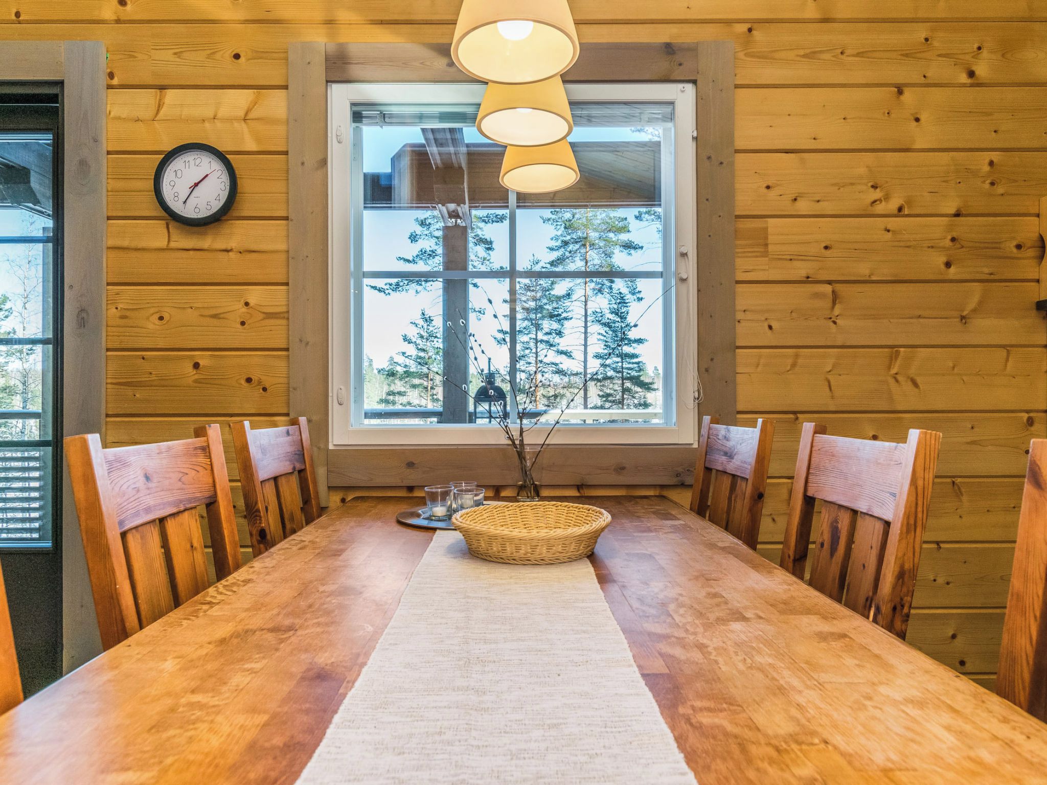 A wooden-paneled room with a dining table, chairs, a clock, and a window with a view of trees.