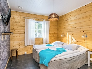 Ein Zimmer mit Holzboden, einem Bett, einem Fenster mit Vorhängen, einem kleinen Tisch und einem an der Wand befestigten Fernseher.
