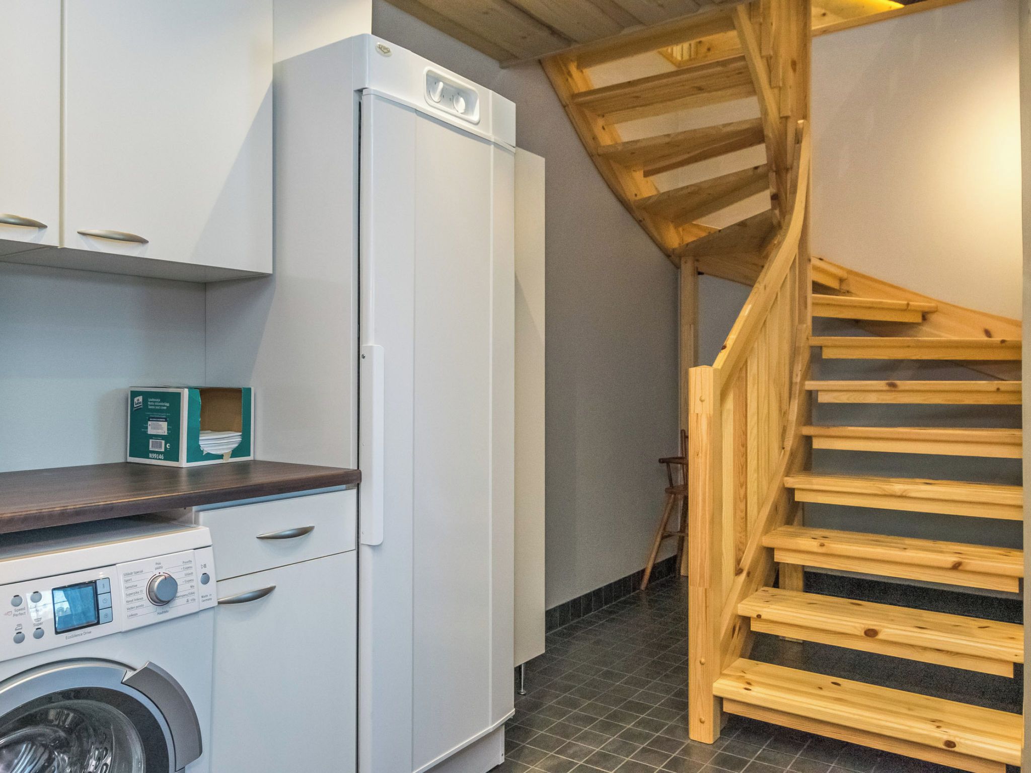 A compact kitchen with a washing machine, a refrigerator, and a wooden staircase.