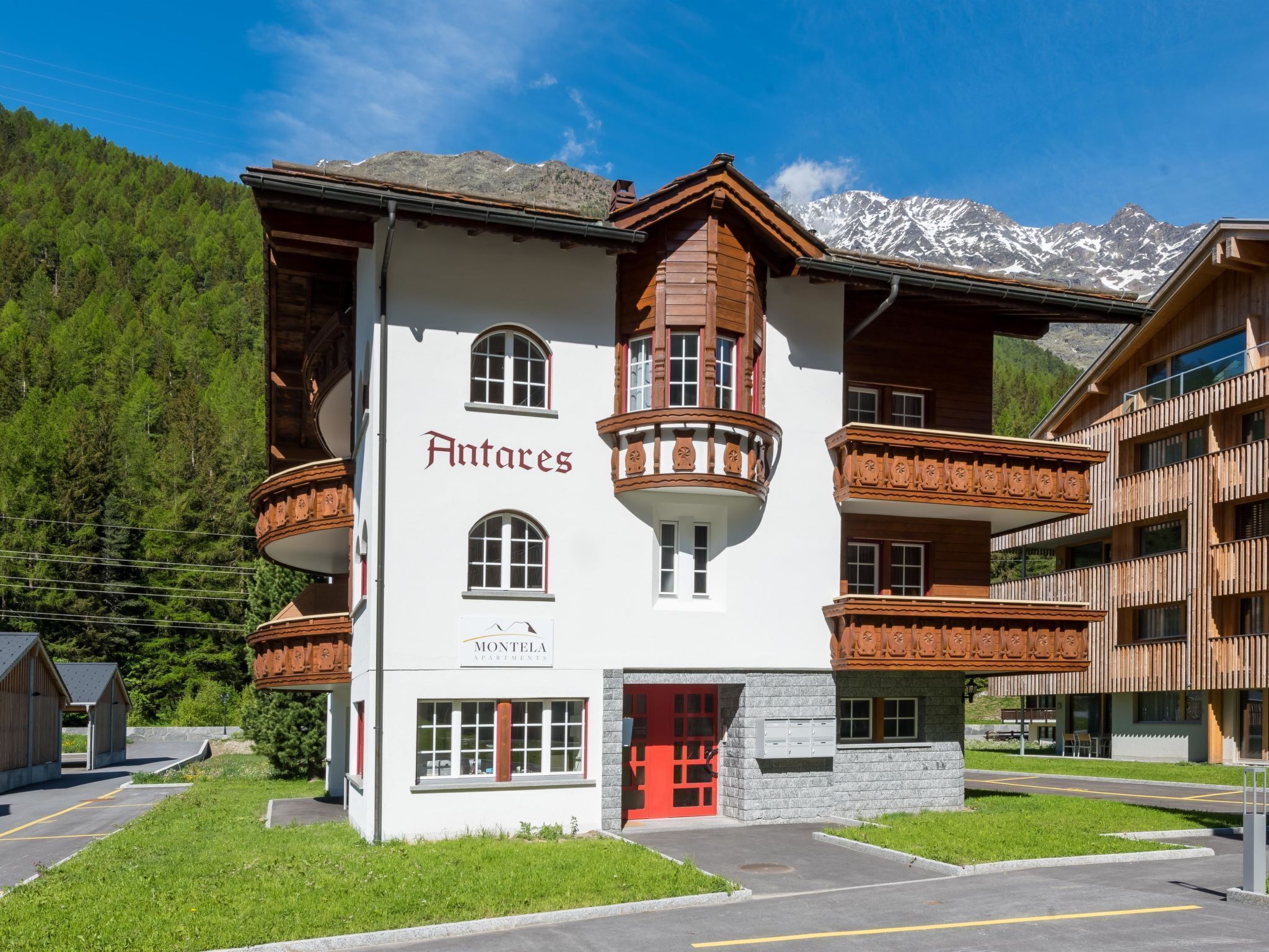 A building with a red door, labeled "Antares Montela Apartments," featuring a traditional architectural style with wooden balconies and a mountainous backdrop.