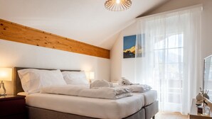 Makuuhuoneessa on suuri sänky, puupalkit katossa, yöpöytä, työpöytä ja ikkuna, jossa on ohuet verhot.