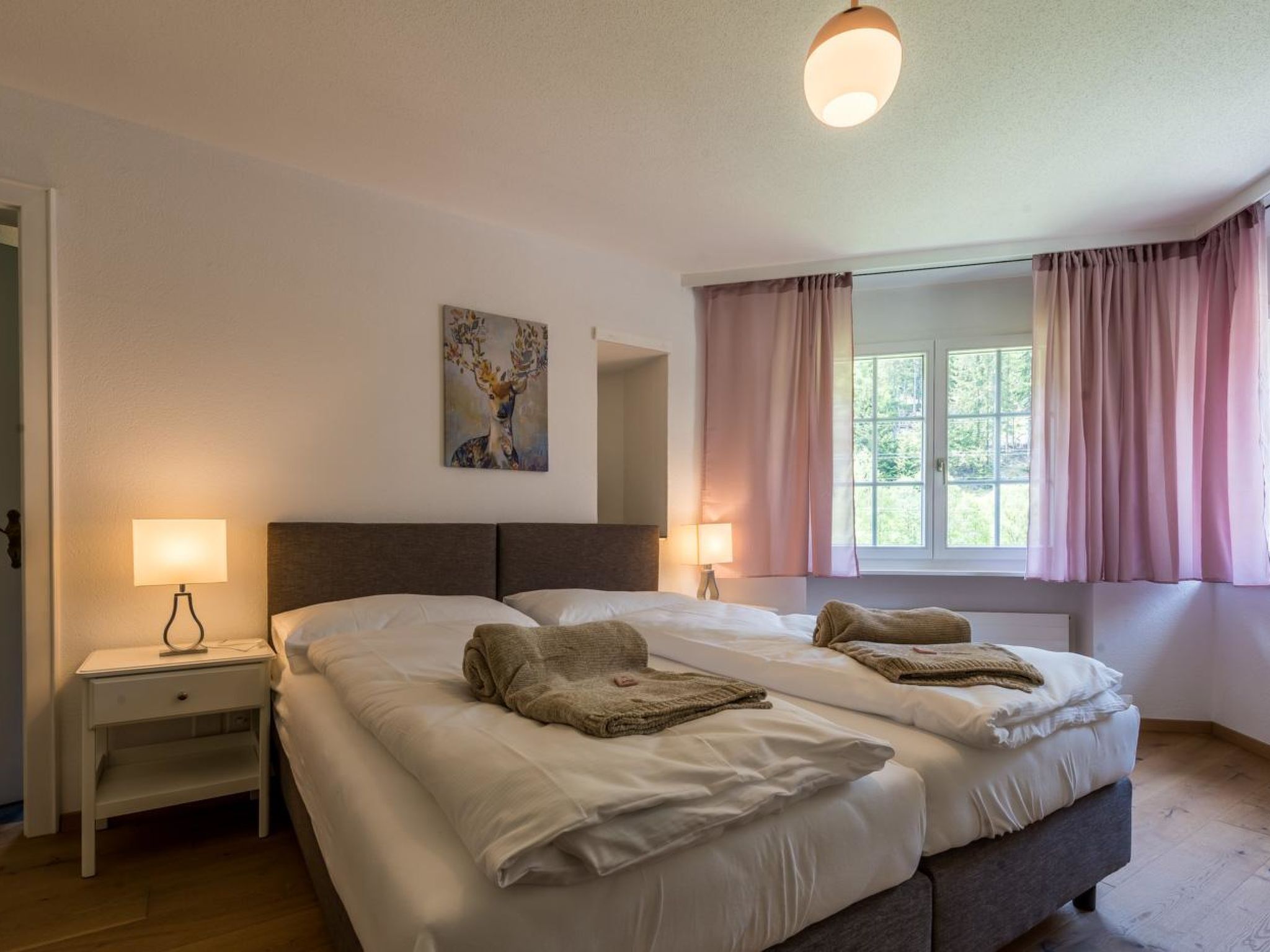 Un letto matrimoniale con due cuscini e una coperta, un comodino con una lampada, un quadro al muro e una finestra con tende.
