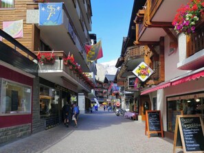 A narrow street lined with shops and restaurants, featuring balconies with flower baskets and flags.