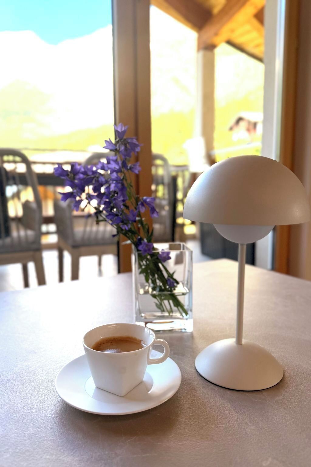 Uma mesa com uma xícara de café, um vaso com flores roxas e um abajur branco.