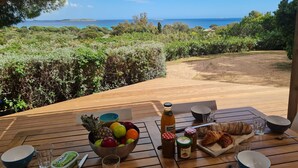 Une table en bois, sur laquelle sont disposés un panier de fruits, des bouteilles et des pâtisseries, se trouve sur une terrasse offrant une vue sur un paysage côtier.