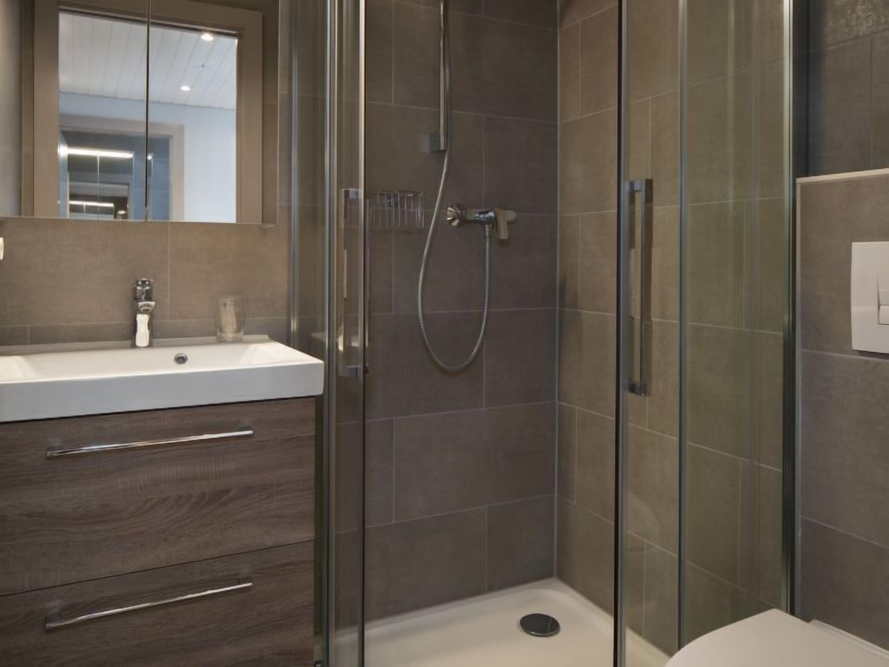 Un baño moderno con ducha envuelta en cristal, lavabo blanco y mueble de tocador de madera oscura con cajones.
