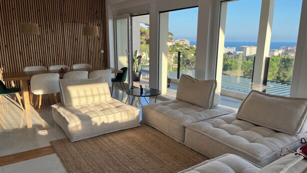 Una sala de estar moderna con un sofá seccional grande, una mesa de comedor y vistas a la ciudad y al mar a través de amplios ventanales.