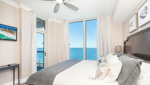 Ein Hotelzimmer mit einem großen Bett, einem Deckenventilator, einem Fernseher und Meerblick durch die Fenster.