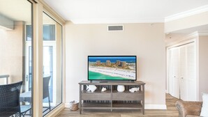 Ein Wohnzimmer mit Fernseher, der eine Strandansicht zeigt, einer grauen Unterhaltungsecke, einem gemusterten Teppich und einer Glastür zum Außenbereich.