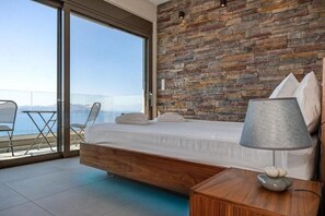 Un dormitorio con una cama grande, una mesita de noche de madera, una lámpara y vista al mar a través de un ventanal.