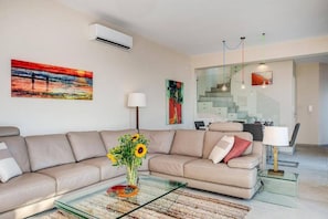 Una sala de estar moderna con un sofá color beige, una mesa de centro de vidrio y un jarrón con girasoles. En la pared hay un cuadro y una escalera que lleva al área de comedor.