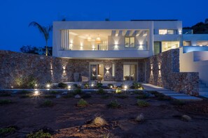 Une maison moderne à deux étages, dotée d’une façade en pierre, de grandes fenêtres et d’un éclairage extérieur.