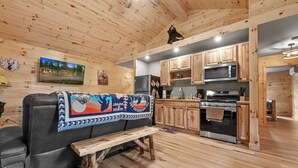 A cozy cabin interior with a wooden ceiling, a black leather sofa with a colorful blanket, a wooden bench, a kitchen with wooden cabinets and appliances, and a mounted picture of a cabin. - Cabin # 3 at Northern Comfort w/opt side by side rentals (Atlanta)