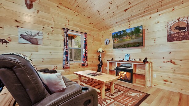 A cozy cabin interior with a leather sofa, a wooden table, a fireplace, and wall art including a moose and a scenic landscape. - Cabin # 3 at Northern Comfort w/opt side by side rentals (Atlanta)