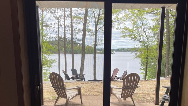 A view through a sliding glass door to an outdoor deck with a fire pit, two Adirondack chairs, and a bench. - Escape to Paradise: Your dream vacation awaits in our stunning lakefront rental! (Tomahawk)