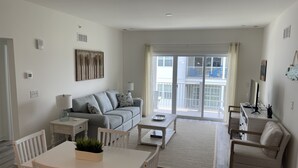 A modern living room with a sofa, a dining table with chairs, a TV, and a balcony with a railing. - Condo on Rehoboth Bay close to beaches and boating (Lewes)