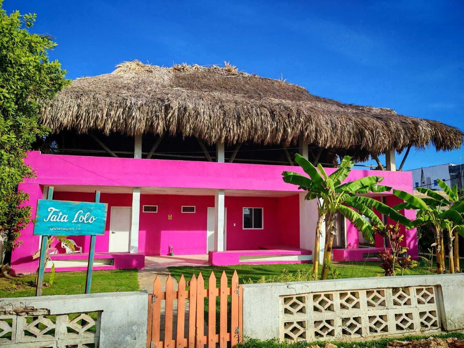Un edificio rosa con techo de paja, un letrero que dice "Tata Lolo" y una cerca de madera al frente.