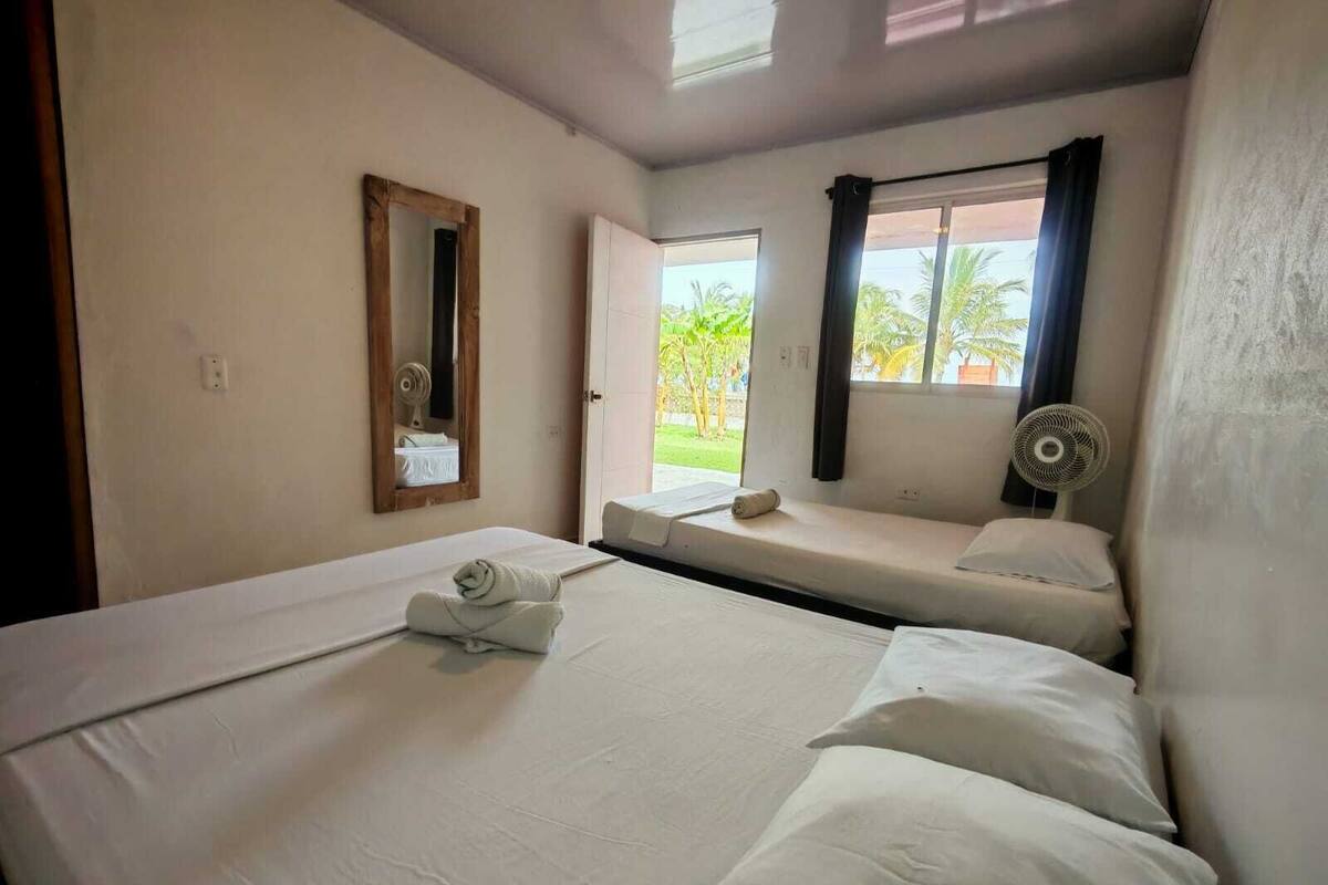 A hotel room with two beds, a window with curtains, a fan, and a mirror on the wall.