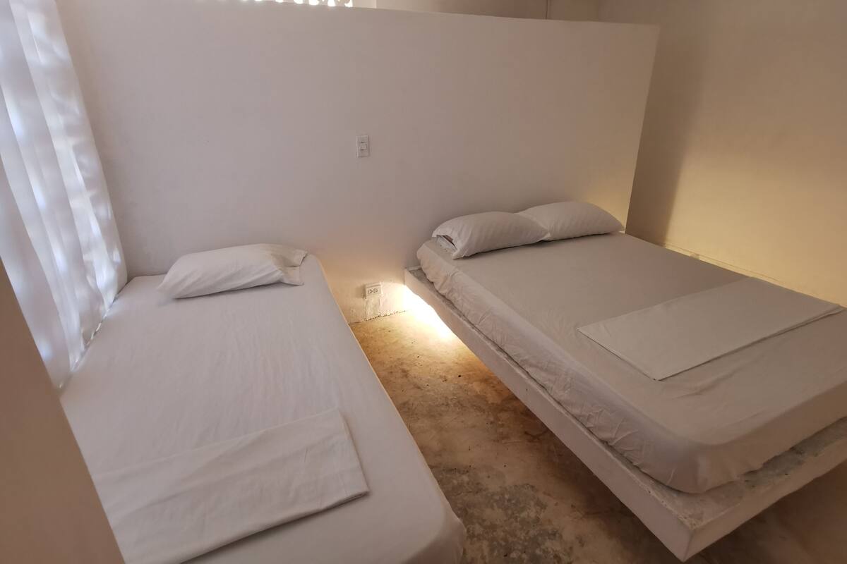 A small, single-room with two single beds, a plain white wall, and a simple window with a curtain.