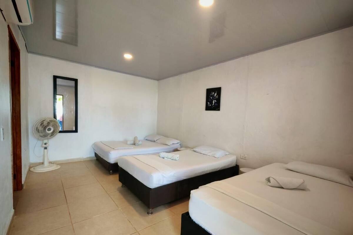 A hotel room with three beds, a standing fan, and a small wall-mounted picture.