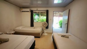Une chambre d’hôtel avec deux lits, une petite table, un appareil de climatisation, une fenêtre donnant sur un espace verdoyant et un ventilateur.