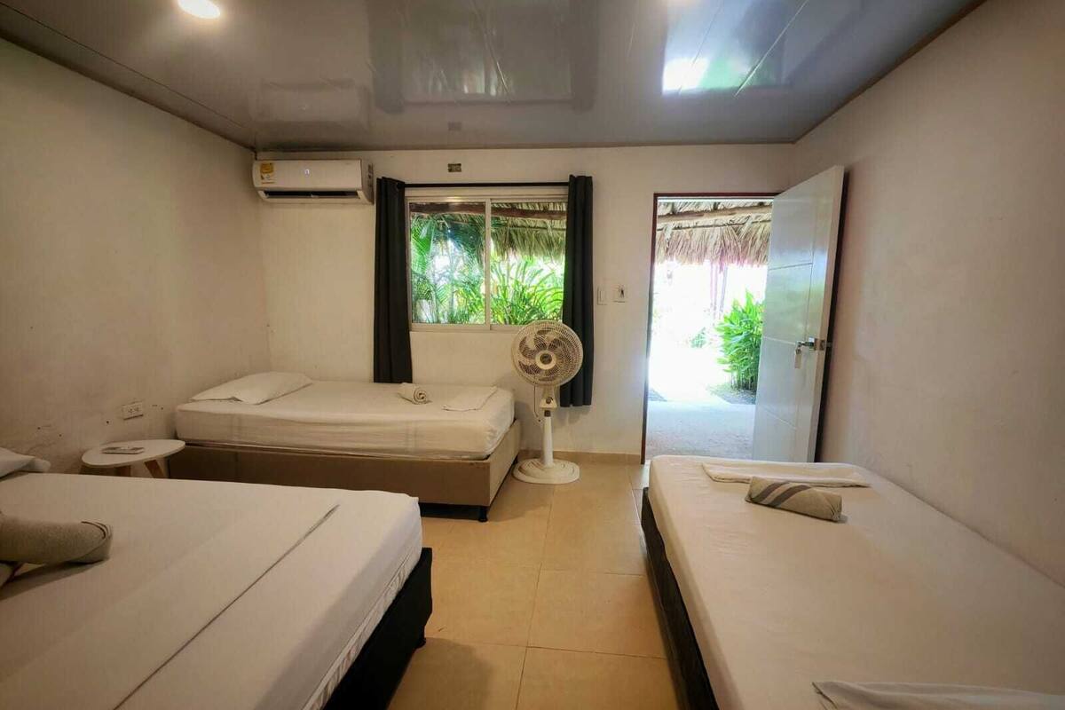 A hotel room with two beds, a small table, an air conditioning unit, a window with a view of greenery, and a fan.