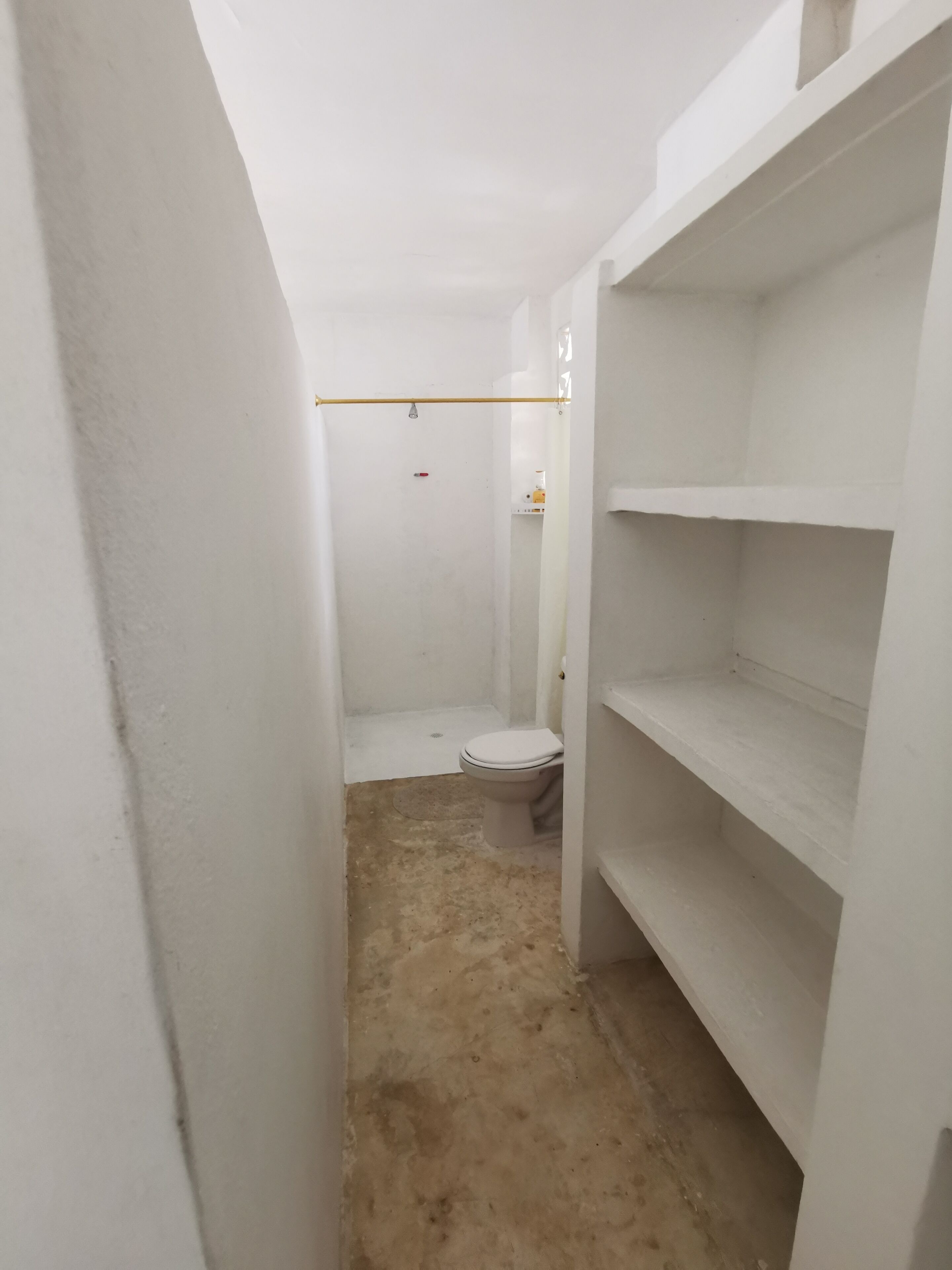Une petite salle de bain aux murs blancs, équipée de toilettes, d’une douche et d’une étagère.