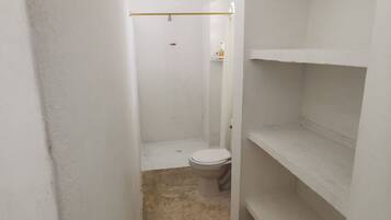 A narrow, white-walled bathroom with a toilet, a shower area, and a shelf unit.