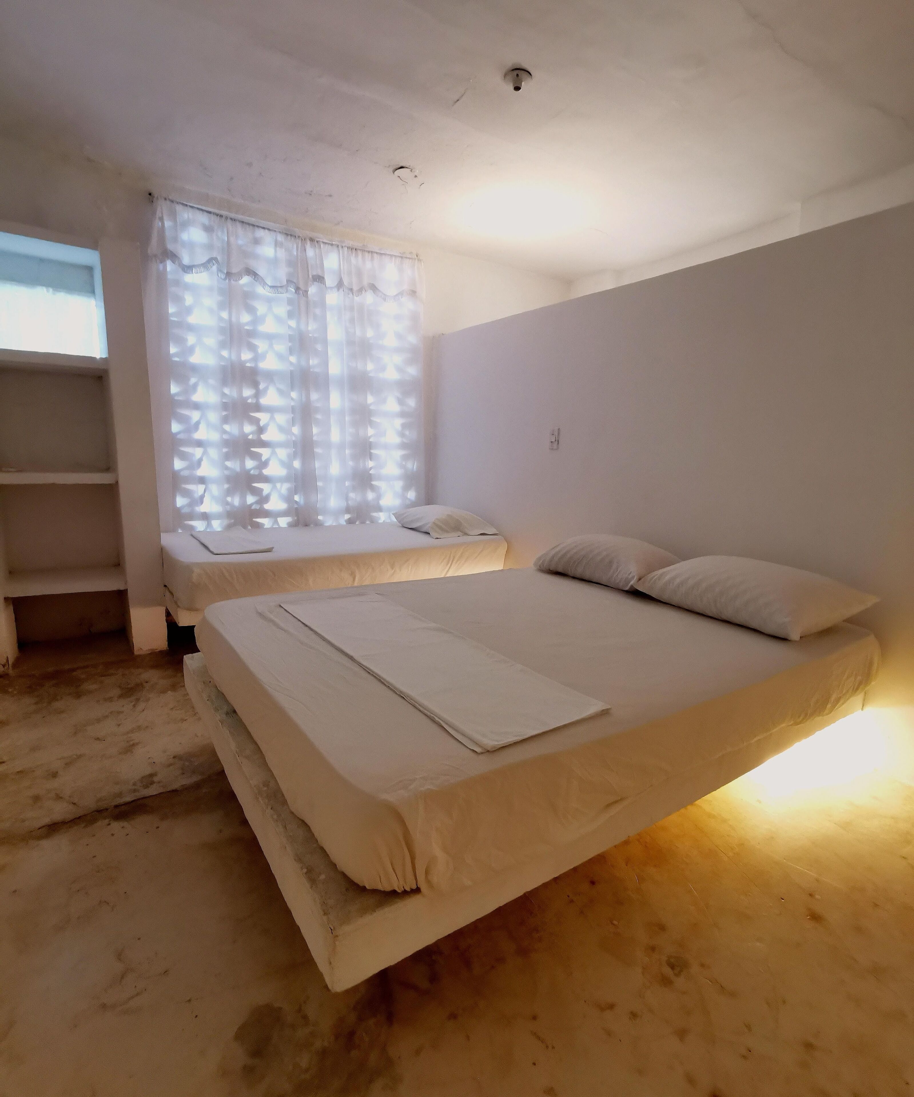 A room with two beds, a window with patterned curtains, and a shelf unit.